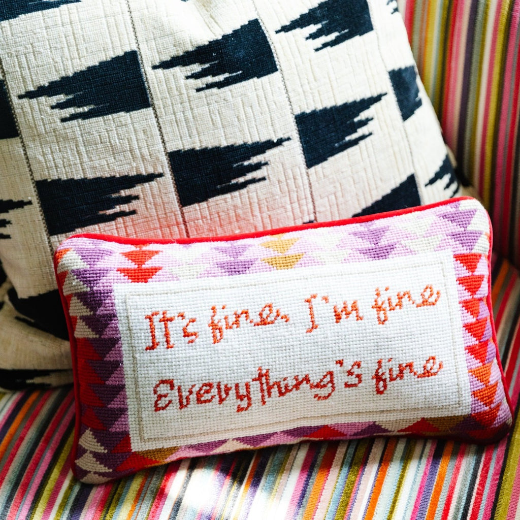 a view of a hand embroidered in wool and backed in luxe lilac velvet chic needlepoint pillow with "It's fine, I'm fine Everything's fine" cheeky saying in front sitting on a colorful couch with a black and white throw pillow at the back