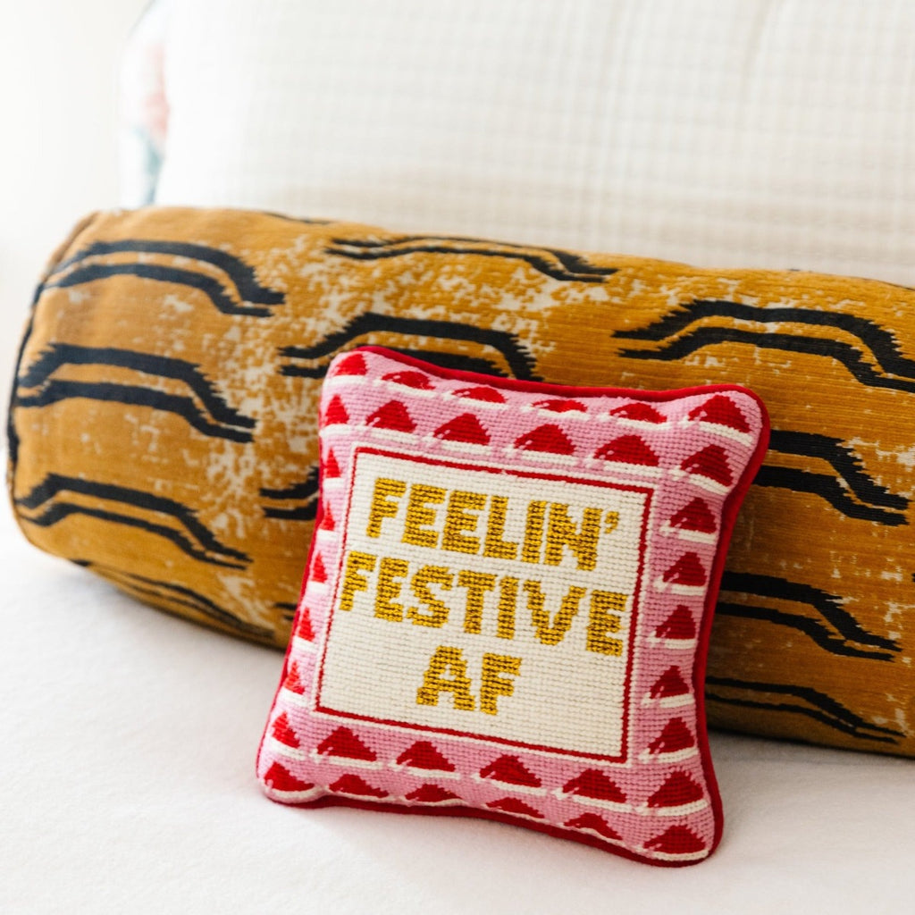 a closer look of a hand embroidered in wool and backed in luxe bright red velvet chic needlepoint pillow with "Feelin' Festive AF" cheeky saying in front sitting on a couch with a yellow bolster pillow at the back of it