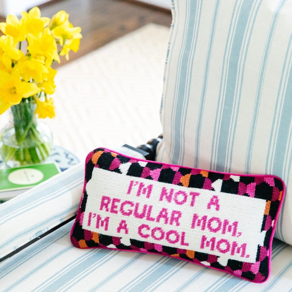 frontal view of a luxe hot pink chic hand needlepoint pillow with "I'm not a regular mom, I'm a cool mom" saying in front while sitting on a chair