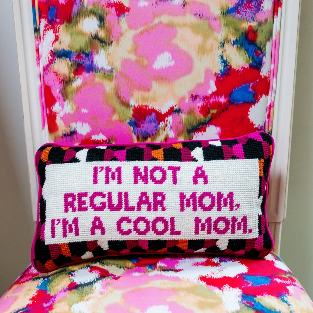 frontal view of a luxe hot pink chic hand needlepoint pillow with "I'm not a regular mom, I'm a cool mom" saying in front while sitting on a colorful chair