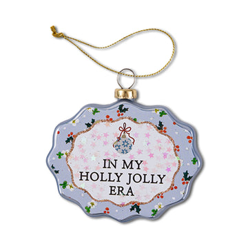 A scalloped glass ornament with a Christmas theme, featuring text 'IN MY HOLLY JOLLY ERA' on a round blue background with red and green accents and a holly design.