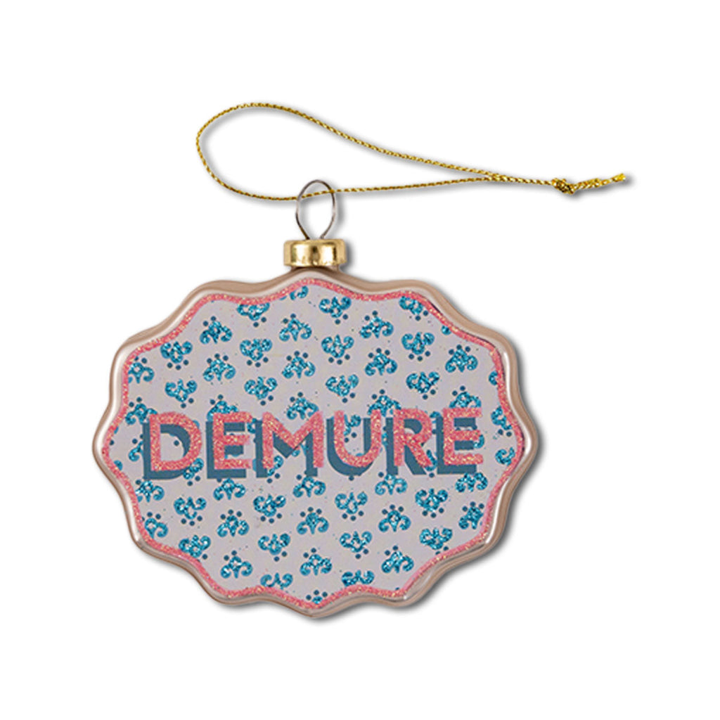 A glass Christmas ornament with a scallop shape, featuring a floral pattern and the word 'DEMURE' displayed prominently in the center.