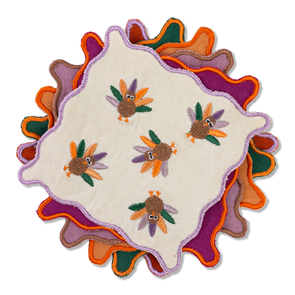 A rectangular cocktail napkin with a scalloped trim featuring block printed Halloween icons such as pumpkins and cats in colors orange, purple, and green on a white background.