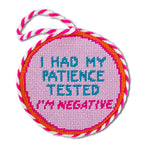 Round pink needlepoint tag with text 'I had my patience tested, I'm negative' on a white background