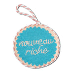 Round needlepoint ornament with 'nouveau riche' text on a white background