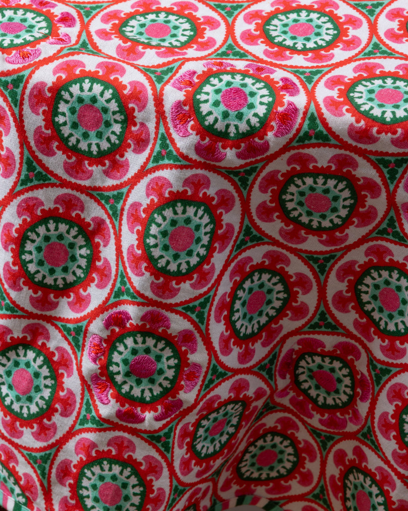 A round, multicolored embroidered tablecloth with geometric patterns, predominantly in red and green shades.