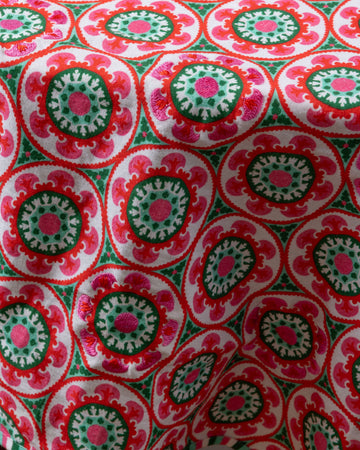 A round, multicolored embroidered tablecloth with geometric patterns, predominantly in red and green shades.