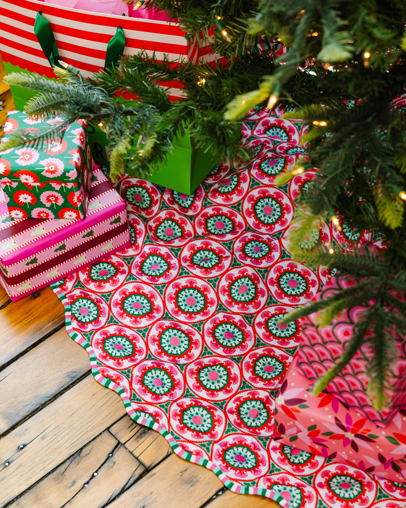 Decorative Christmas tree skirt with red and green pattern on a wooden floor with gifts underneath a tree.