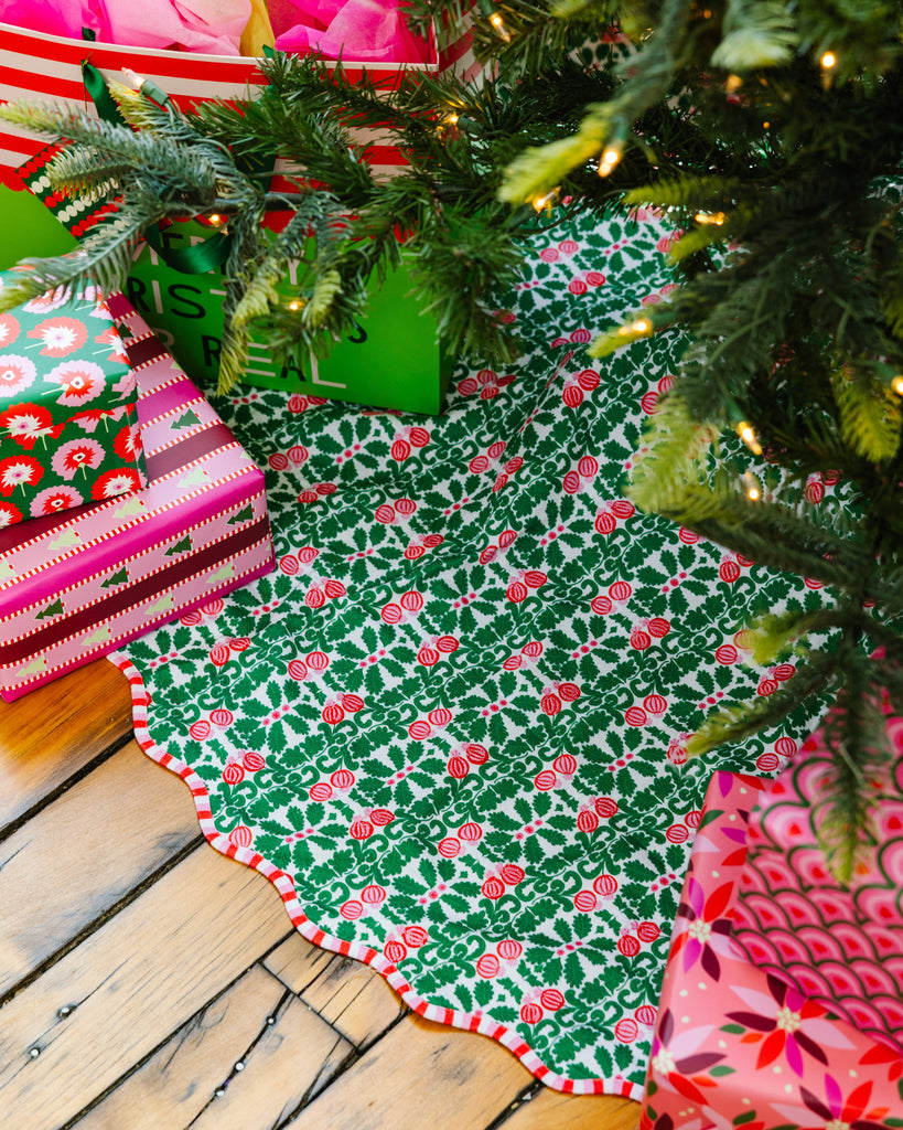 Decorative Christmas tree skirt with holly pattern on a wooden floor next to wrapped gifts.