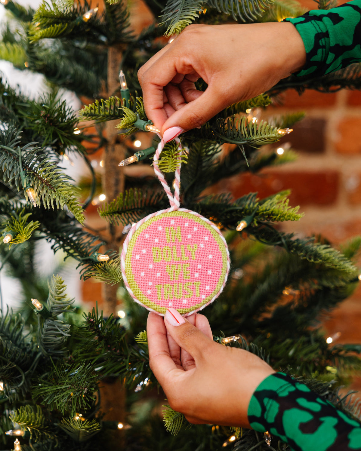 Person hanging a pink and green ornament on a Christmas tree with a brick wall background.