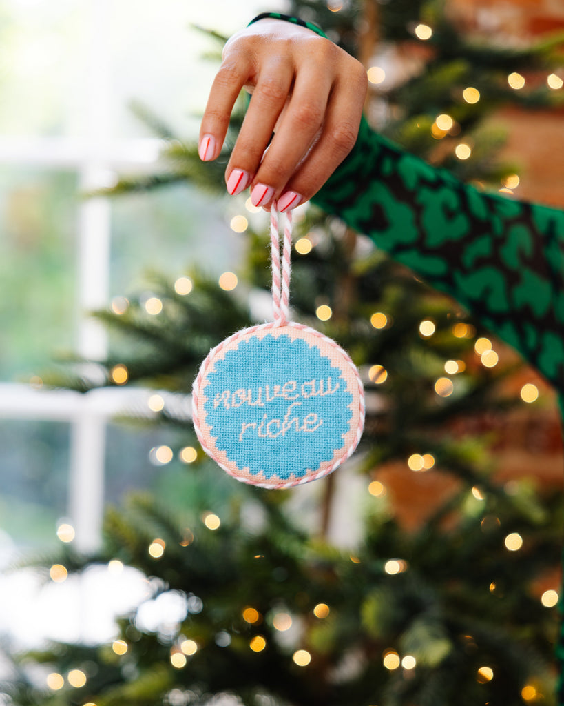 Hand holding a Christmas ornament with 'nouveau riche' text against a decorated tree background