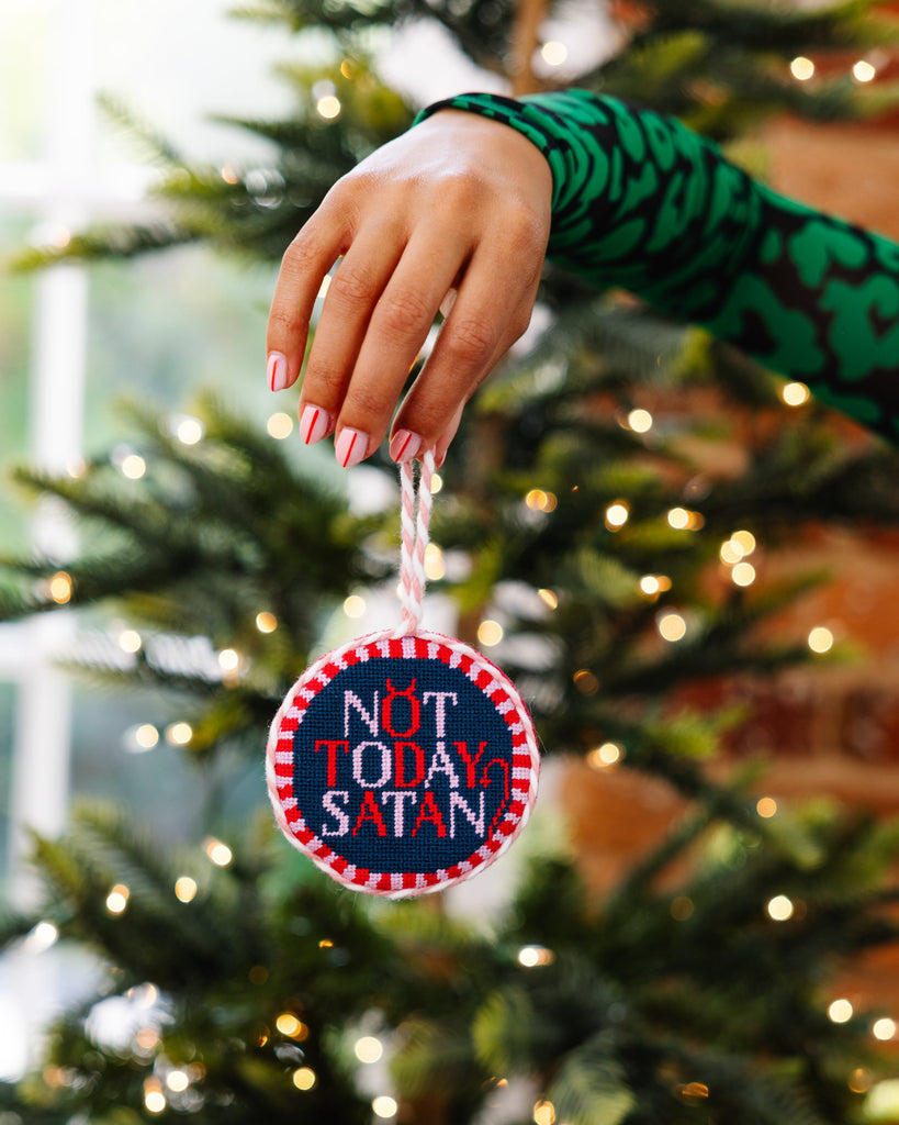 Hand holding a 'Not Today Satan' ornament in front of a decorated Christmas tree.
