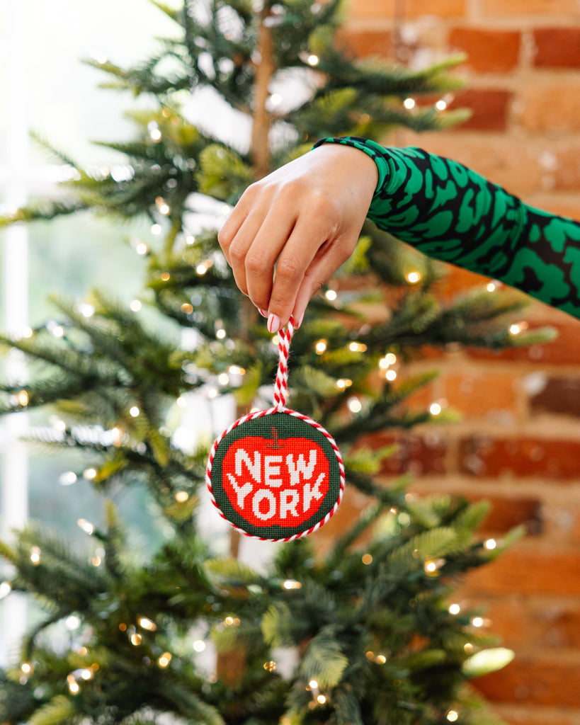 Hand holding a 'New York' ornament in front of a Christmas tree with a brick wall background.