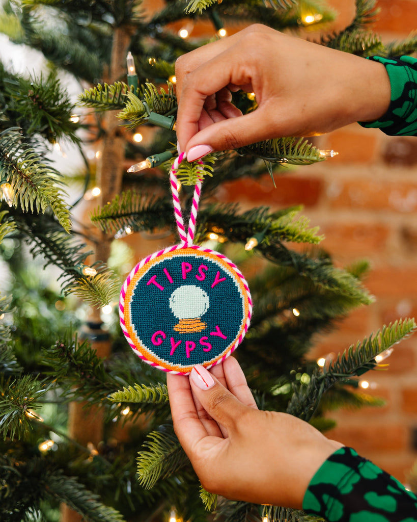 Hand holding a 'Tipsy Gypsy' ornament in front of a decorated Christmas tree.