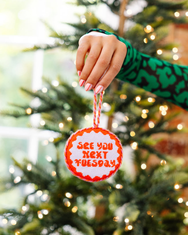 Hand holding a Christmas ornament with 'See you next Tuesday' text against a decorated tree background