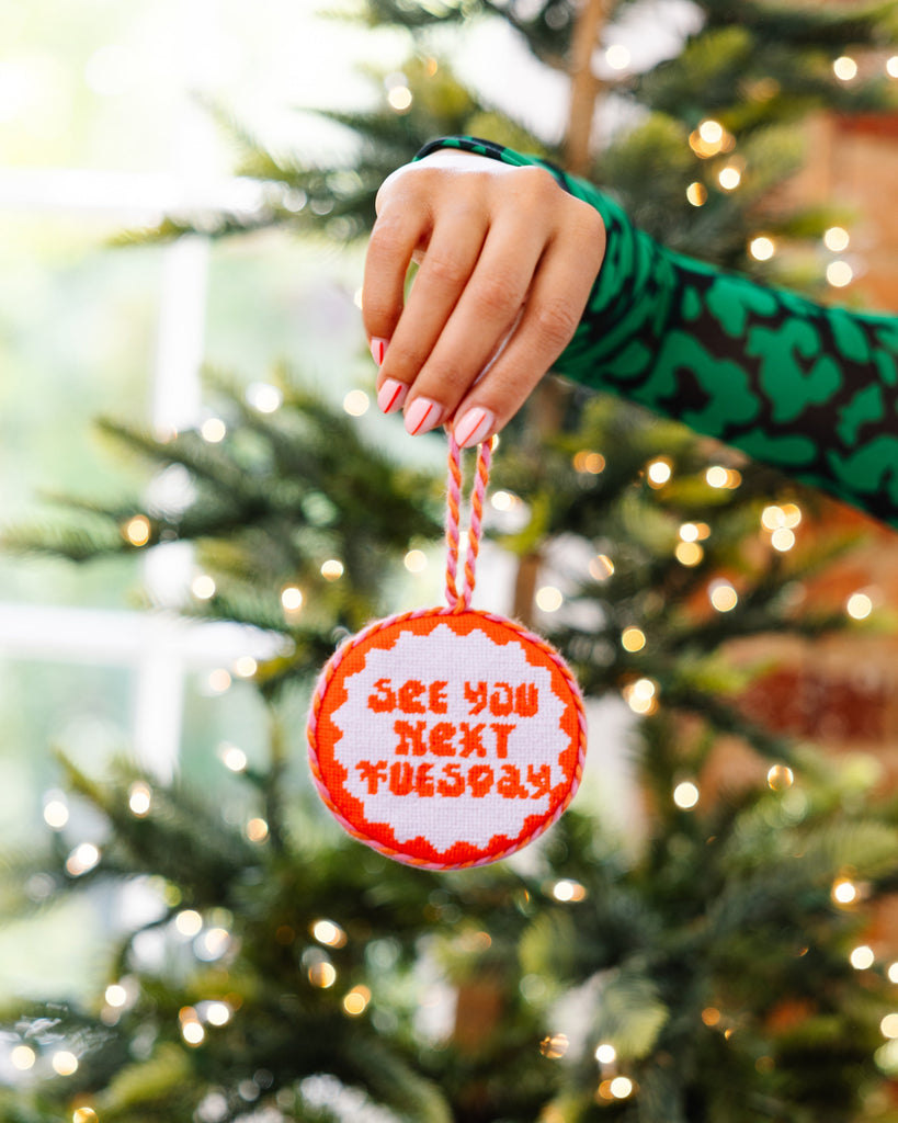 Hand holding a Christmas ornament with 'See you next Tuesday' text against a decorated tree background