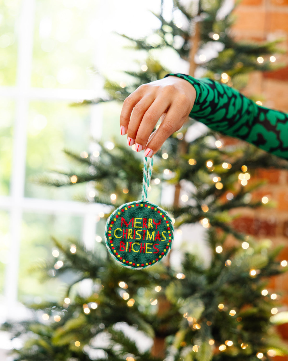 Hand holding a 'Merry Christmas Bitch' ornament in front of a decorated Christmas tree.