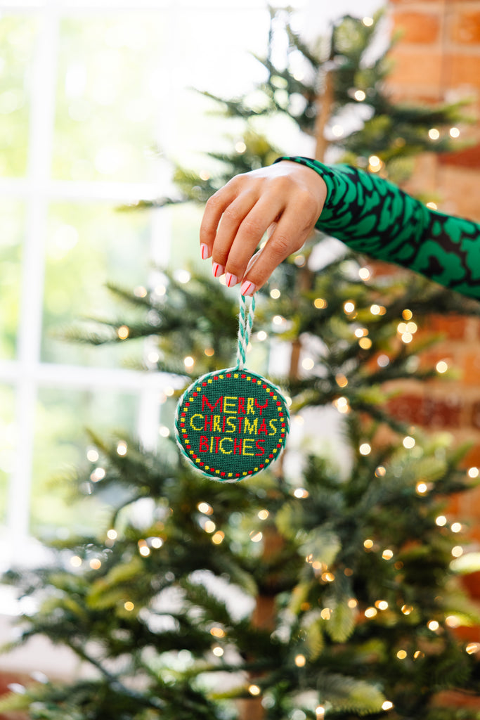 Hand holding a 'Merry Christmas Bitch' ornament in front of a decorated Christmas tree.