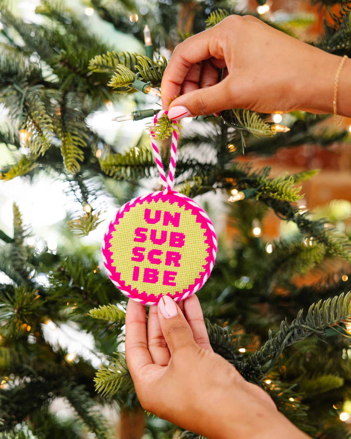 Hand holding a pink and yellow ornament with 'UN SUBSCRIBE' text against a Christmas tree background