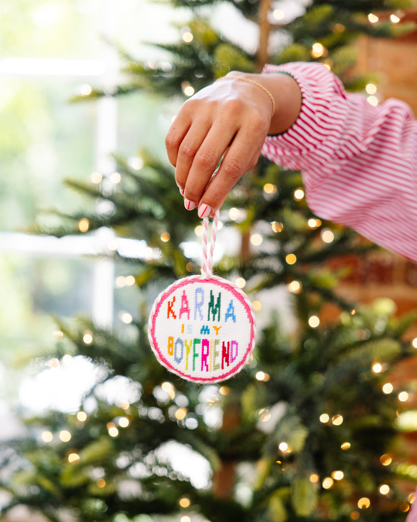 Hand holding a Christmas ornament with 'Karma is my boyfriend' text in front of a decorated tree.