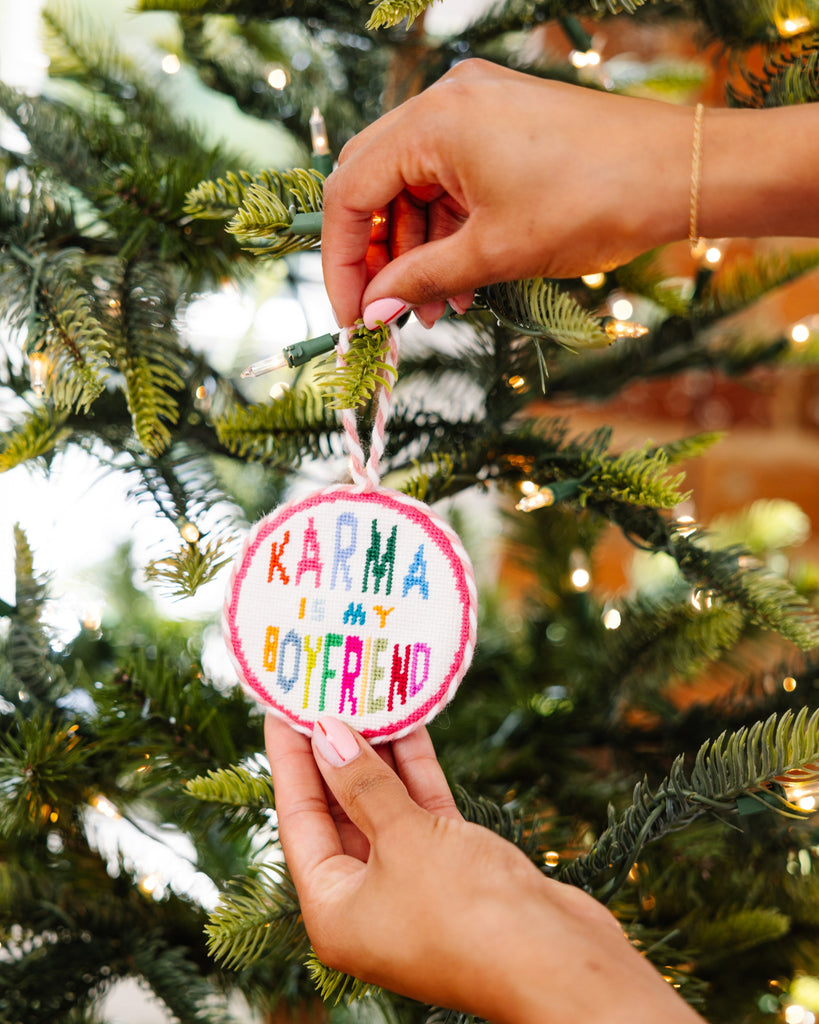 Person hanging a ornament with 'Karma is my boyfriend' text on a decorated Christmas tree.