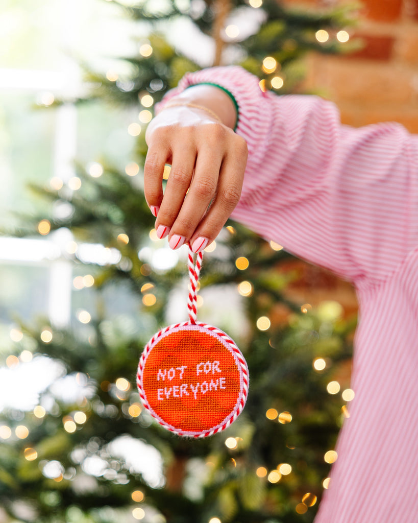 Person holding a Christmas ornament with 'Not for Everyone' text in front of a decorated tree.