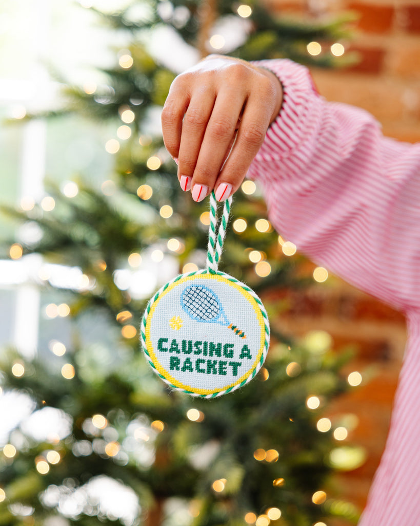 Hand holding a Christmas ornament with 'Causing a Racket' text in front of a decorated tree.