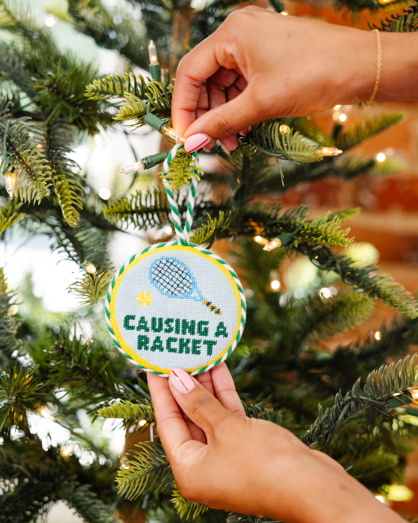 Hand holding a Christmas ornament with 'Causing a Racket' text against a decorated tree background