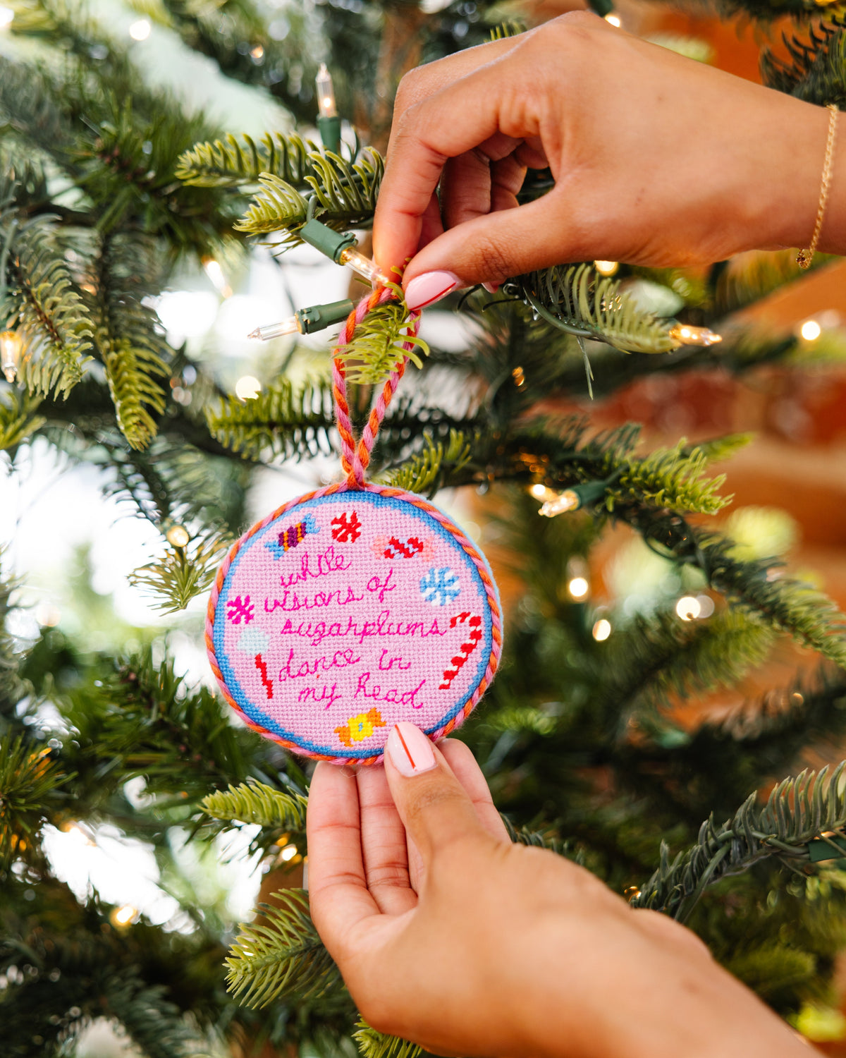 Hand holding a pink embroidered ornament with text against a Christmas tree background