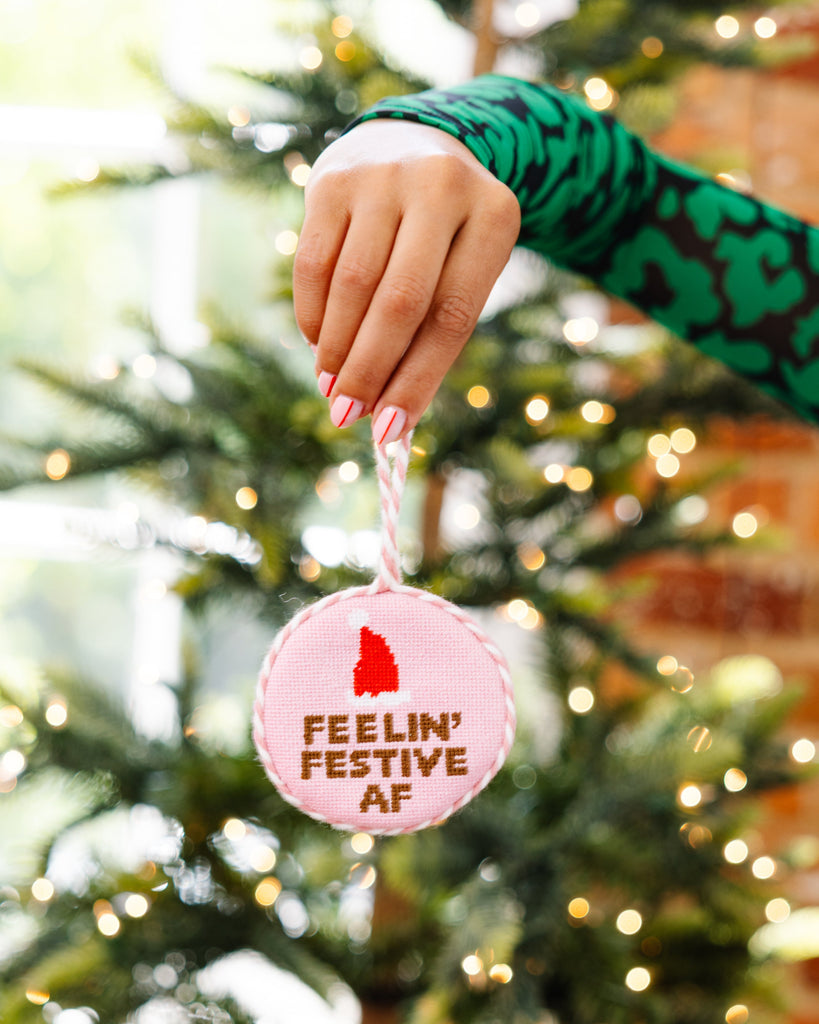 Hand holding a Christmas ornament with 'Feelin' Festive AF' text in front of a decorated tree.