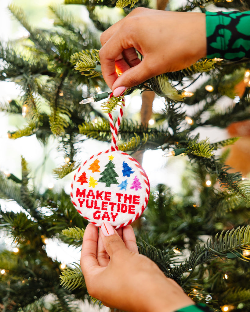 Hand holding a Christmas ornament with 'Make the Yuletide Gay' text on a decorated tree.