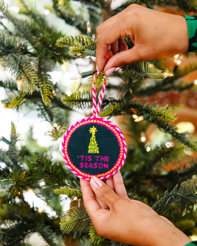 Hand holding a embroidered ornament with 'Tis the Season' text against a Christmas tree background.