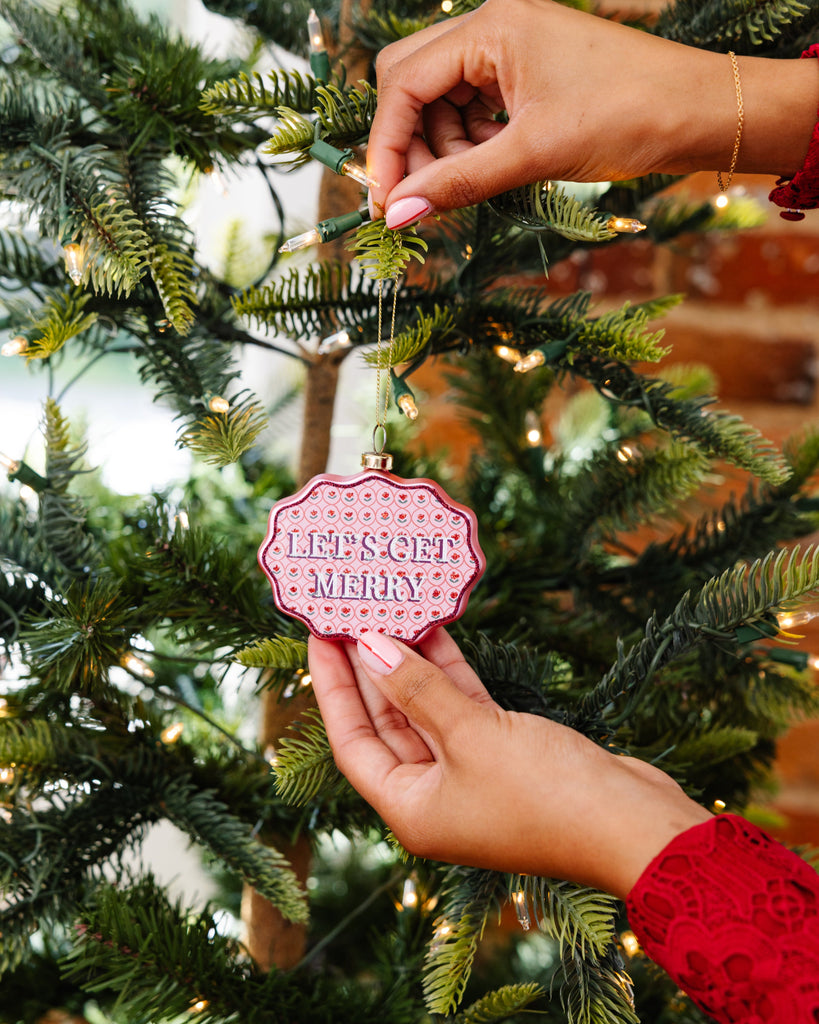 Person hanging a pink ornament with 'Let's Get Merry' text on a Christmas tree.