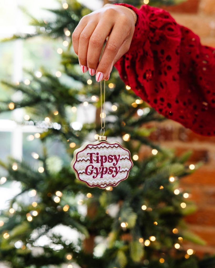 Hand holding a 'Tipsy Gypsy' ornament in front of a decorated Christmas tree.