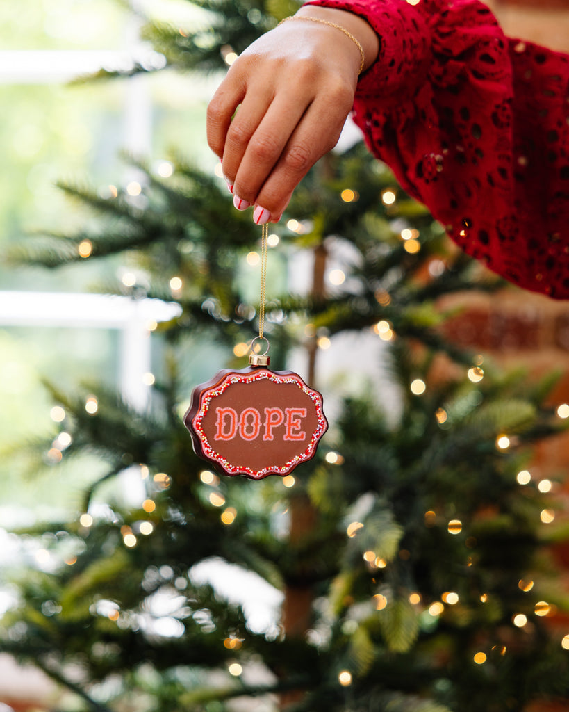 Hand hanging a Christmas ornament with 'DOPE' text in front of a decorated tree.