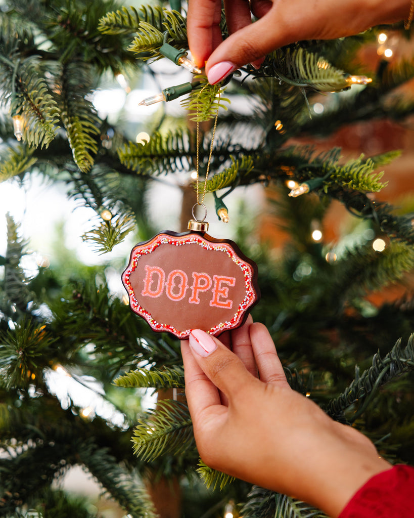Person holding a Christmas ornament with 'DOPE' text on a decorated tree