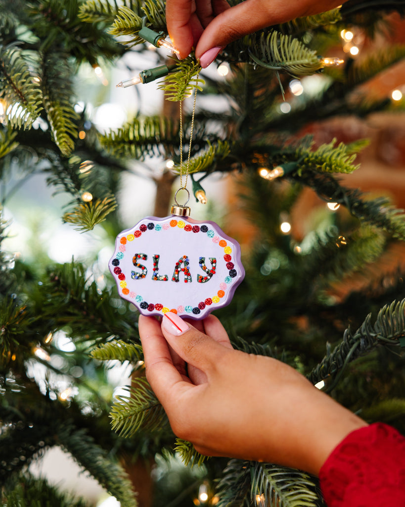 Hand holding a Christmas ornament with 'SLAY' text on a decorated tree