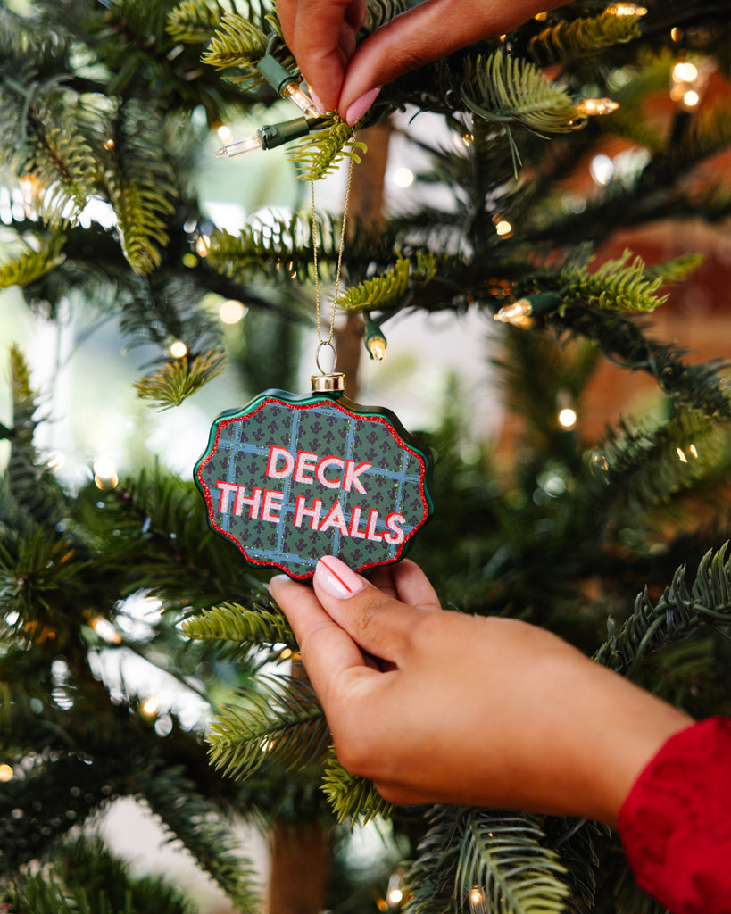 Person hanging a Christmas ornament with 'Deck the Halls' text on a decorated tree.