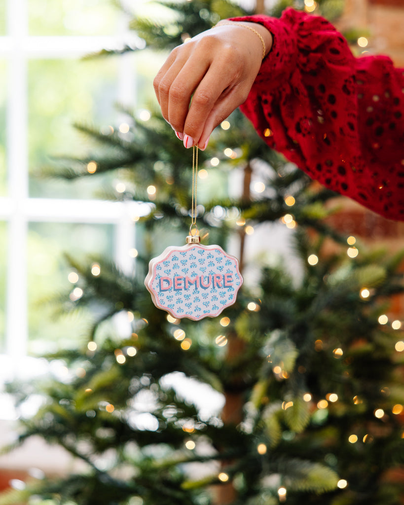 Hand holding a decorative ornament with 'Demure' text in front of a Christmas tree.