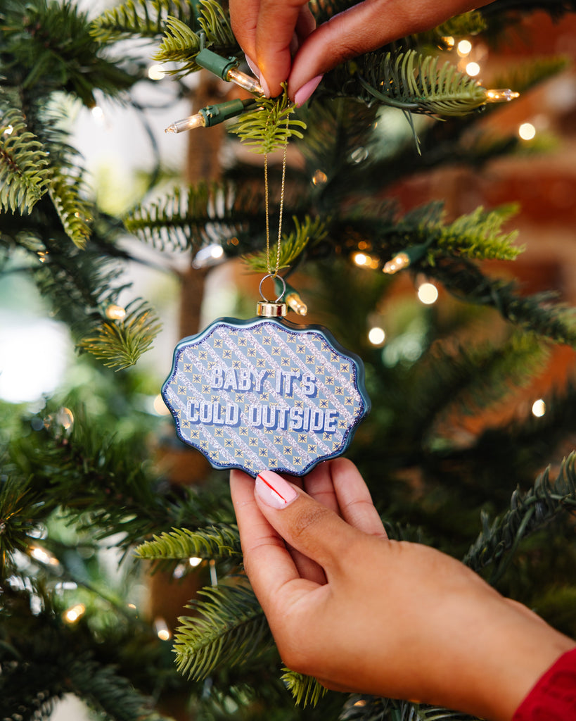 Person holding a Christmas ornament with 'Baby it's cold outside' text on a decorated tree.