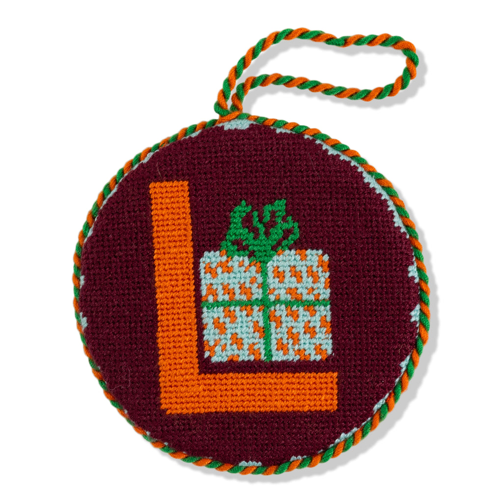 Round needlepoint ornament with letter 'L' and gift design on a white background