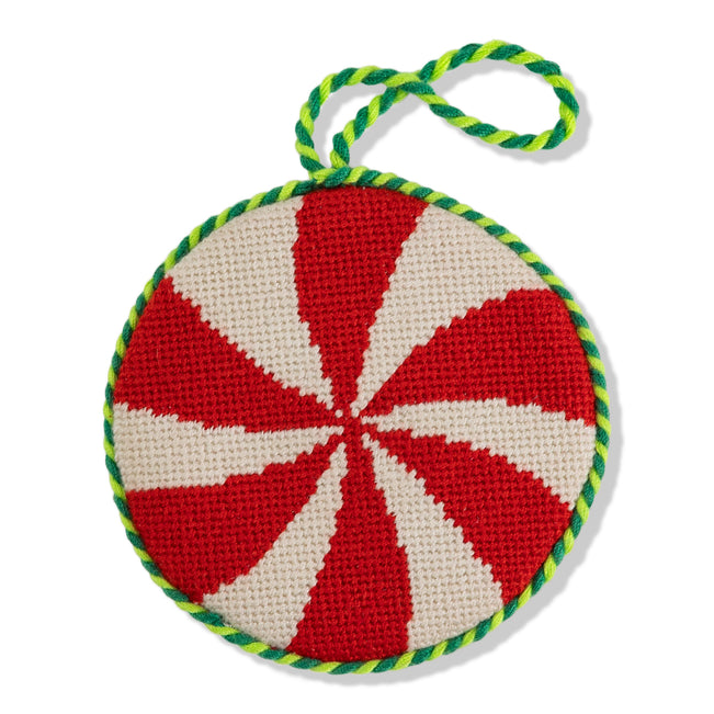 A round needlepoint ornament with a peppermint design, featuring red and white stripes, and a green, white, and red braided edge.
