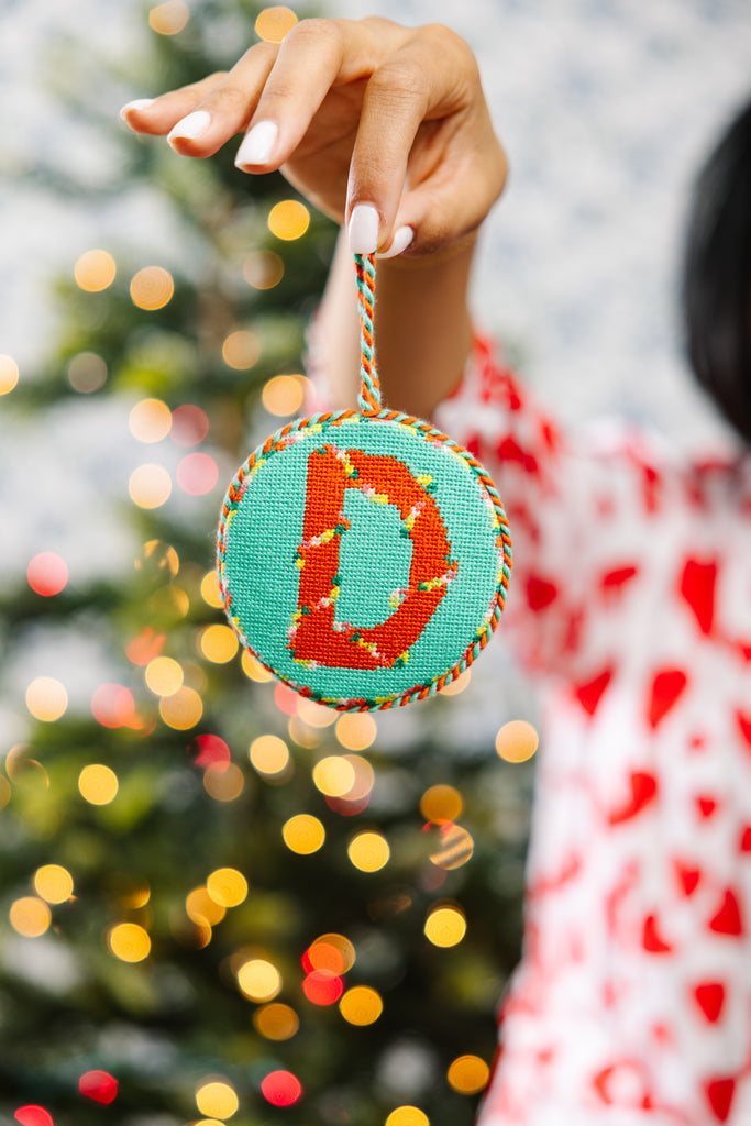 Hand holding a Christmas ornament with a letter 'D' against a blurred Christmas tree background