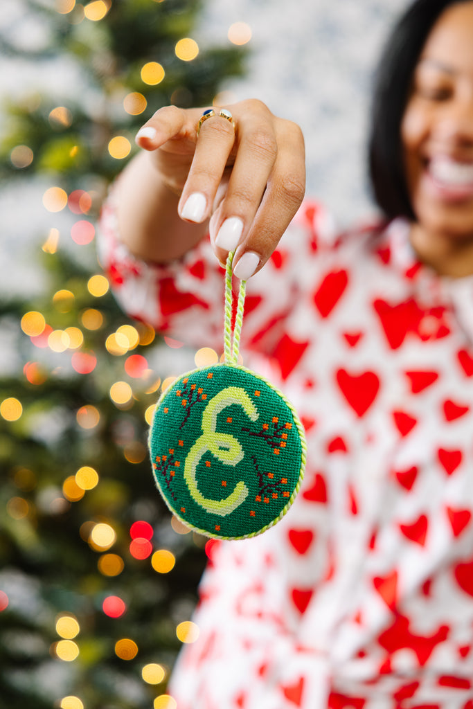 Person holding a green ornament with a letter 'E' in front of a blurred Christmas tree.