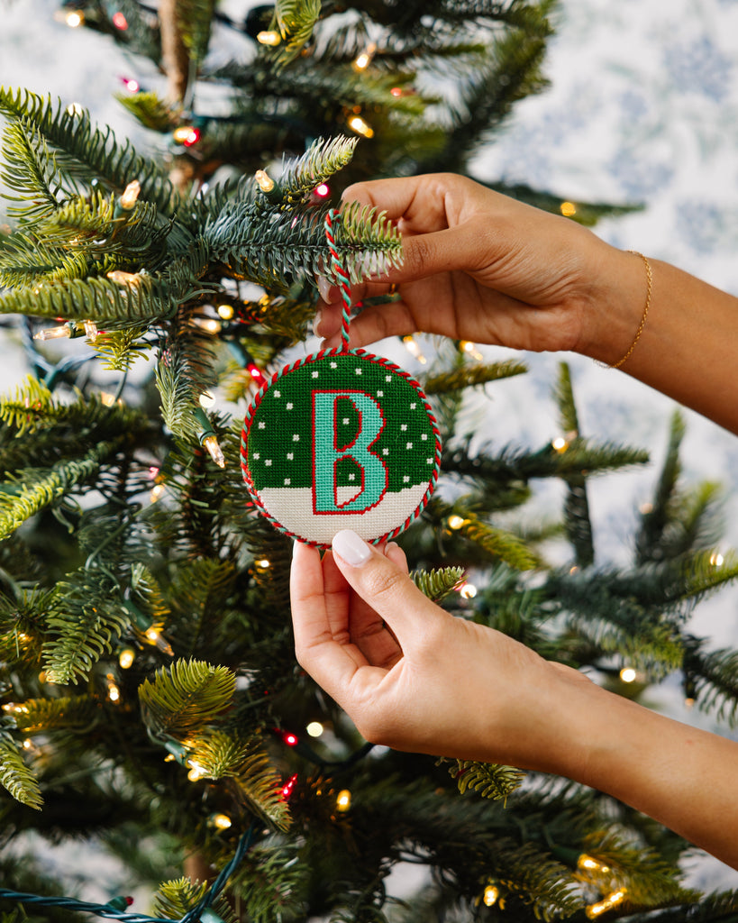 Hand holding a Christmas ornament with a letter 'B' in front of a decorated tree.