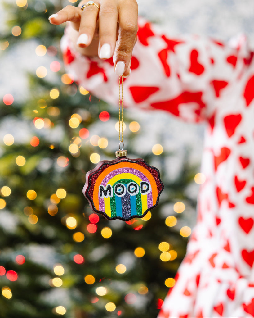 Hand holding a rainbow-colored ornament with 'MOOD' text against a Christmas tree background.