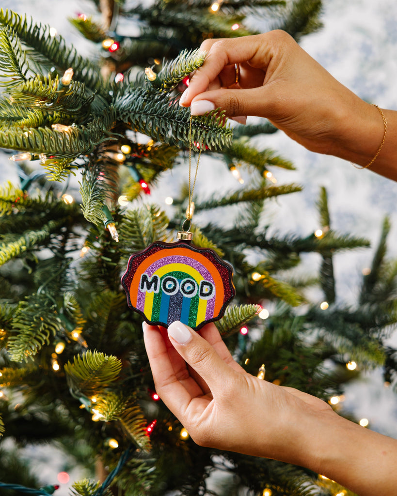 Hand holding a rainbow 'MOOD' ornament against a decorated Christmas tree.