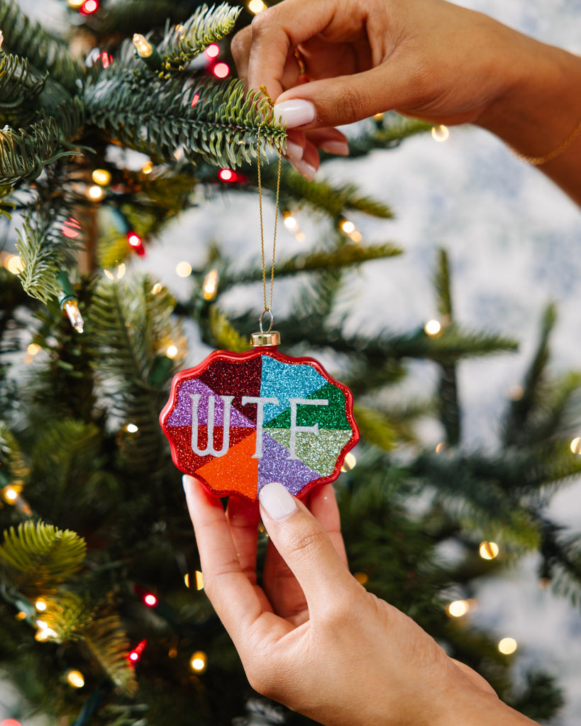 Person holding a colorful ornament with 'WTF?' on a decorated Christmas tree.