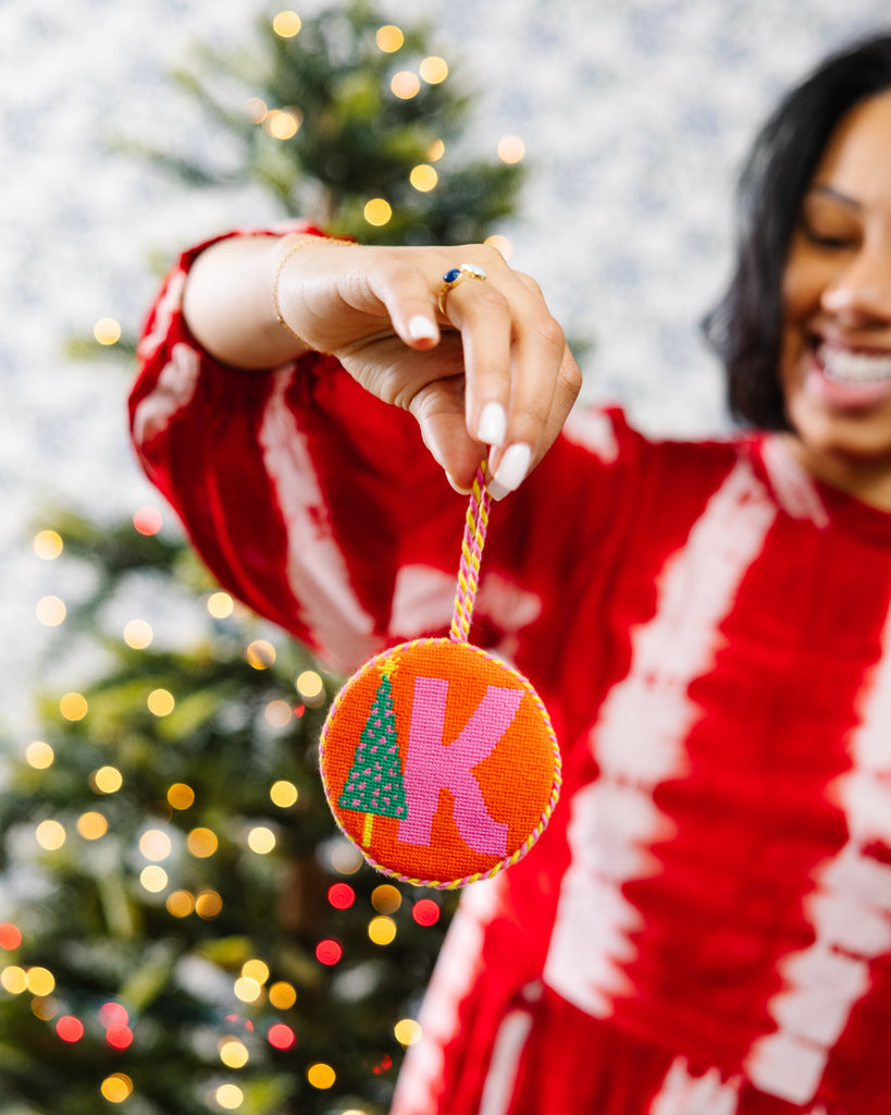 Woman holding a Christmas ornament with a tree and 'K' in front of a decorated tree.