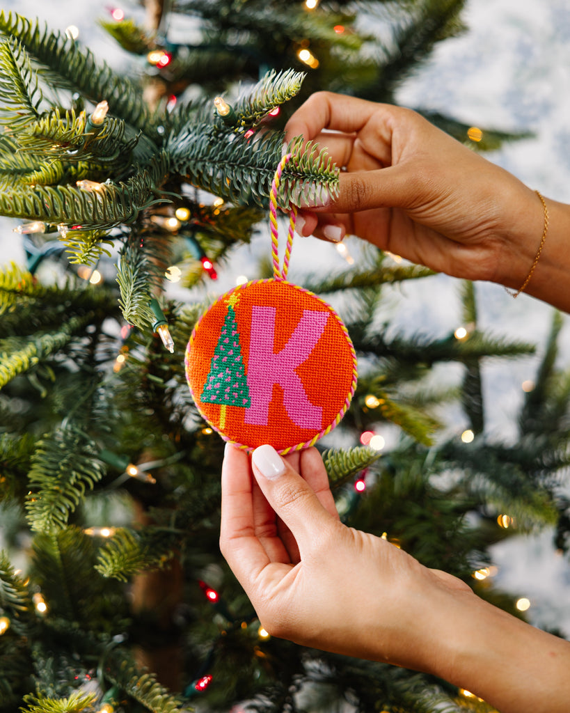 Hand holding a colorful Christmas ornament with letters 'K' and 'A' in front of a decorated tree.